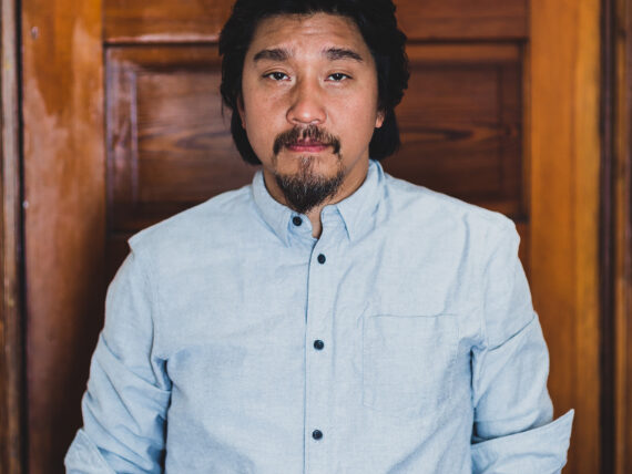 An asian man with a beard wearing a blue button up shirt standing in front of a wood door.