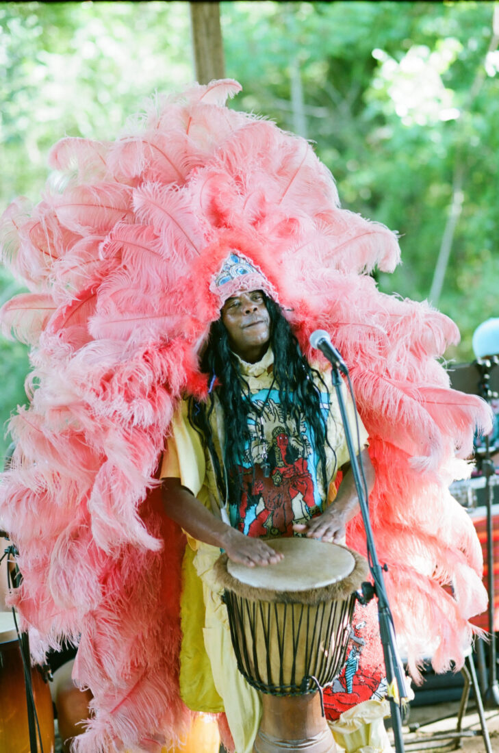 A man plays the drums while wearing a large pink feathered hat