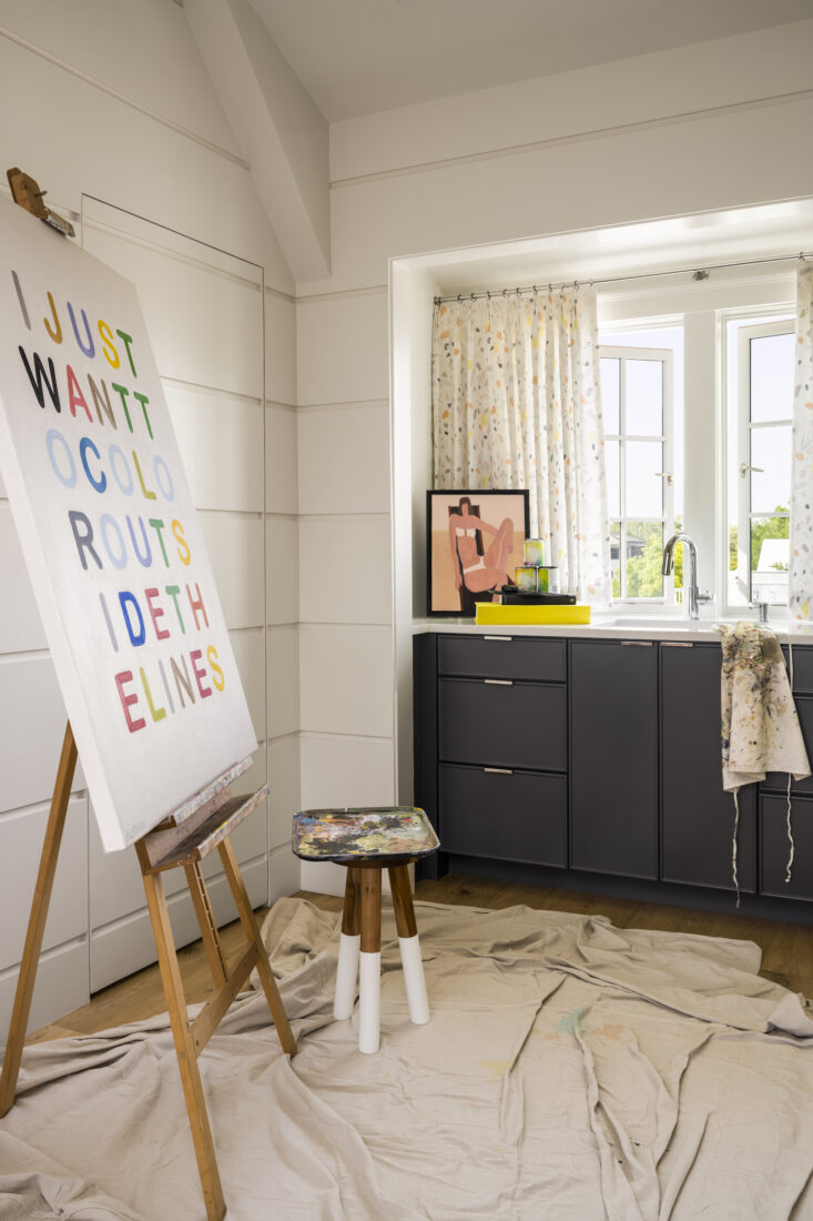 Inside a room with a painting on an easel, a paint palette on a stand, and a sink area with open windows