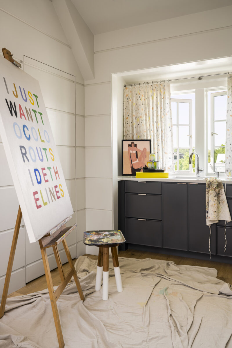 Inside a room with a painting on an easel, a paint palette on a stand, and a sink area with open windows