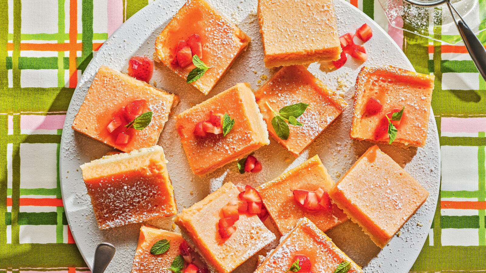 A plate of lemon bars on a plaid background with a bowl of sugar and strawberries