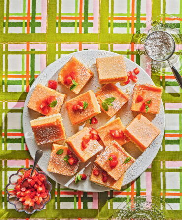 A plate of lemon bars on a plaid background with a bowl of sugar and strawberries