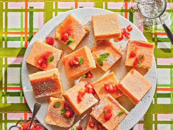 A plate of lemon bars on a plaid background with a bowl of sugar and strawberries