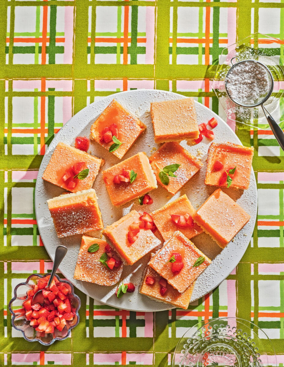 A plate of lemon bars on a plaid background with a bowl of sugar and strawberries