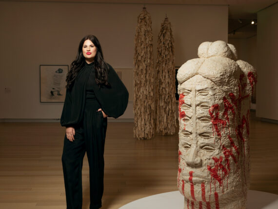 A woman in a black suit stands in a gallery next to a totemic head sculpture with many eyes and red glaze that says "Raven Halfmoon" on the side