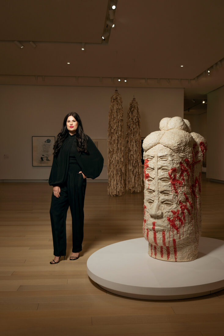 A woman in a black suit stands in a gallery next to a totemic head sculpture with many eyes and red glaze that says "Raven Halfmoon" on the side