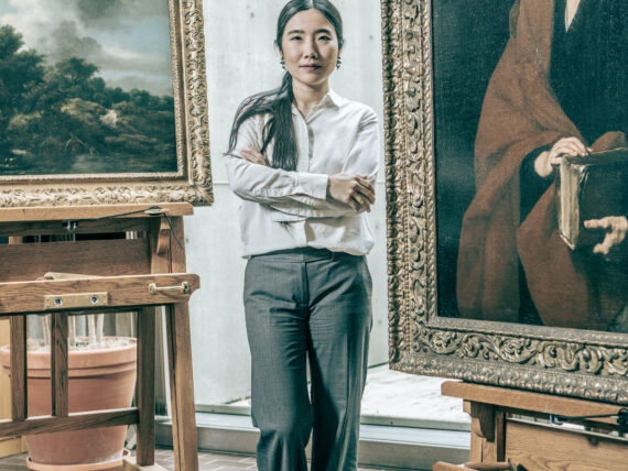 A woman stands in an art conservation studio with a large painting on an easel behind her