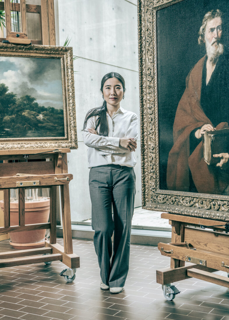 A woman stands in an art conservation studio with a large painting on an easel behind her