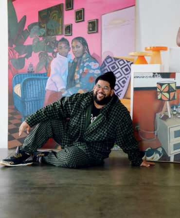 An artist sits on the floor of his studio with a painting of a little girl and a woman behind him