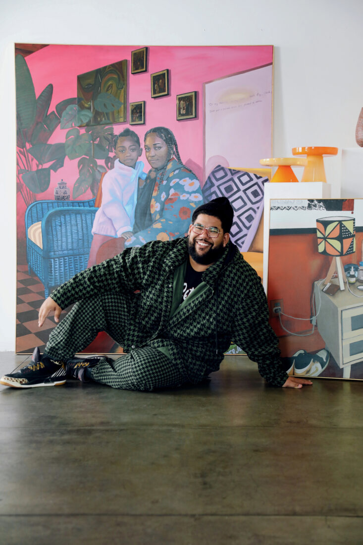 An artist sits on the floor of his studio with a painting of a little girl and a woman behind him