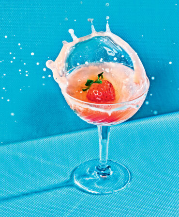 A pink drink splashes as a strawberry goes into it. The glass stands against a blue background