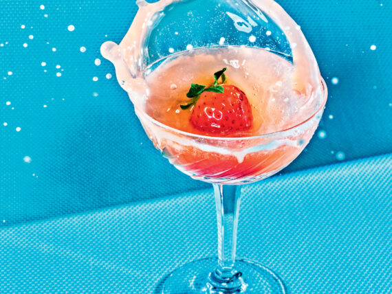 A pink drink splashes as a strawberry goes into it. The glass stands against a blue background
