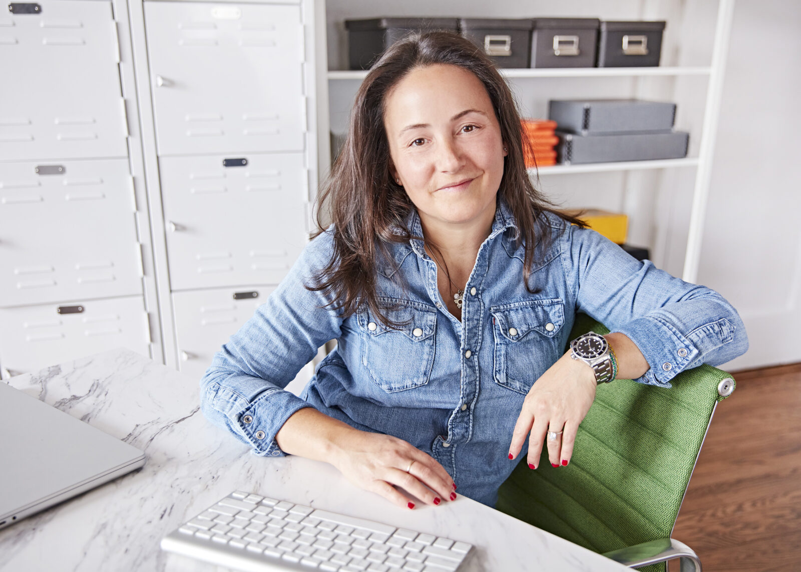 A woman wearing a denim shirt sitting on a green chair at a white desk.