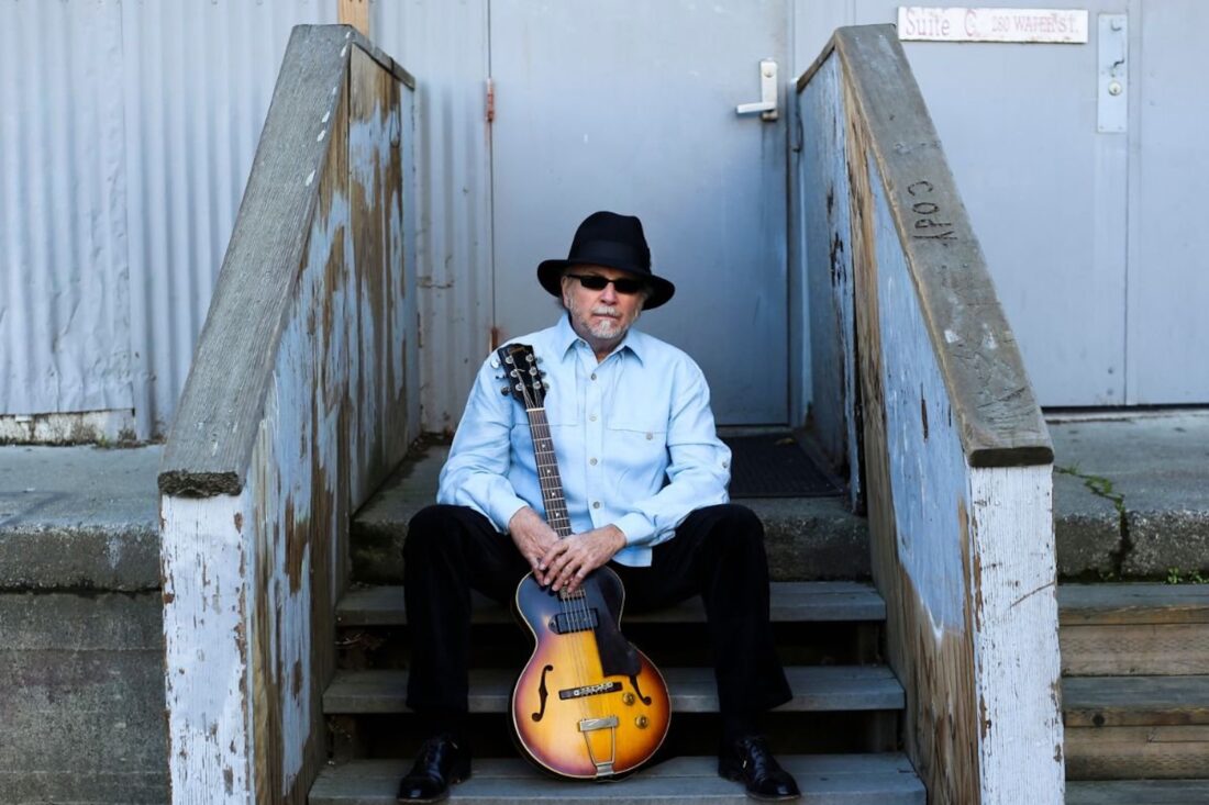 A man in a blue shirt holds a guitar and sits on a porch