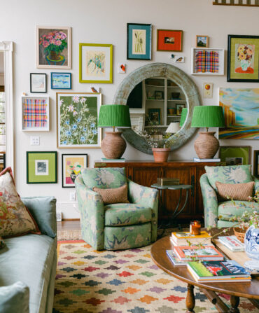 A colorful living room with many works of art on the wall, green chairs, and a patterned rug