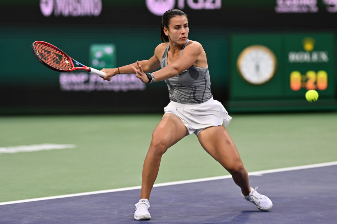 A woman wearing a grey tank top and white skirt playing tennis.