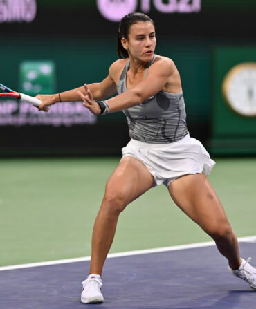 A woman wearing a grey tank top and white skirt playing tennis.