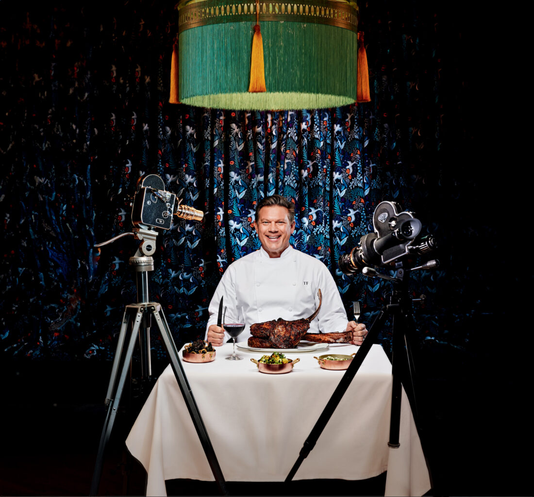 A man sits at a table with food with cameras pointed at him.