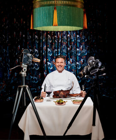 A man sits at a table with food with cameras pointed at him.
