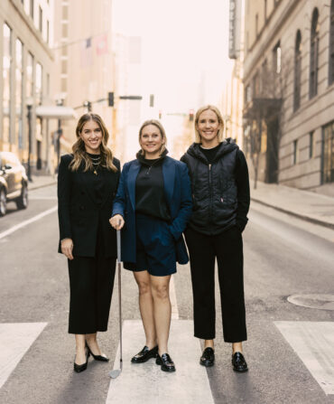 Three women in stylish clothing stand in a street and smile
