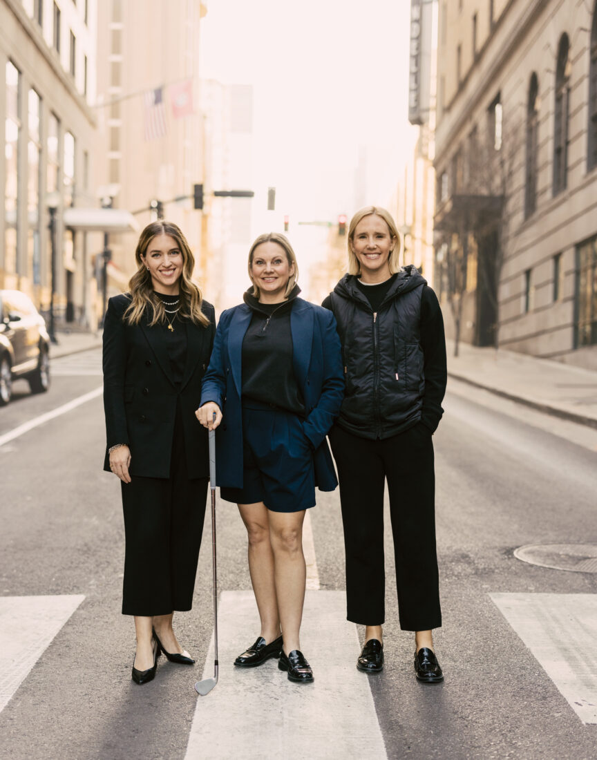 Three women in stylish clothing stand in a street and smile