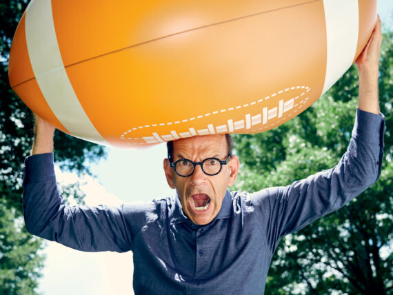 A man holds a giant football above his head with his mouth open in a yell