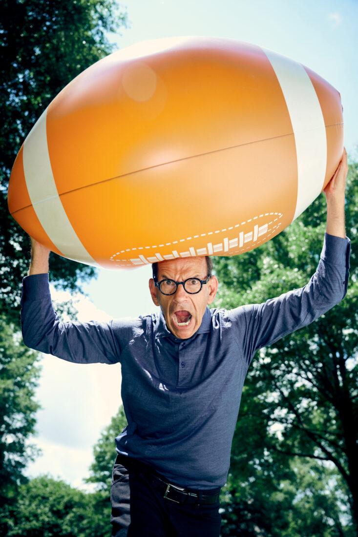 A man holds a giant football above his head with his mouth open in a yell