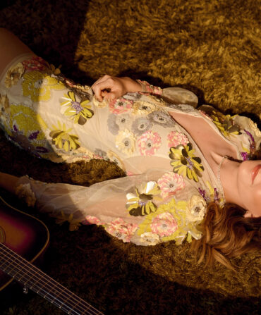 A woman lays in a fuzzy carpet with a guitar. She wears a sheer dress with green and purple flower appliques.