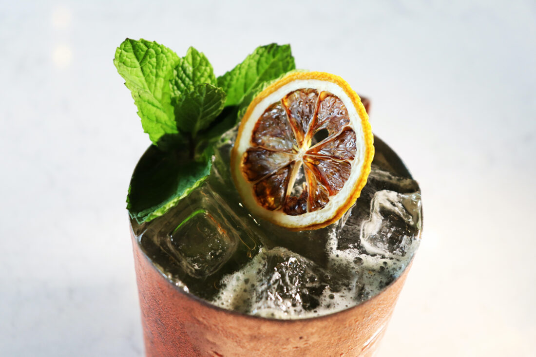 A cocktail in a copper cup, garnished with a lemon and mint