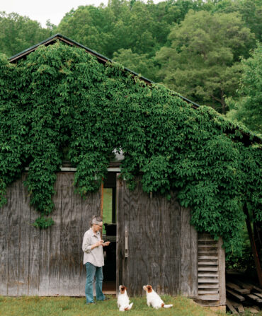 A woman stands in front of a vine-covered barn with two small dogs