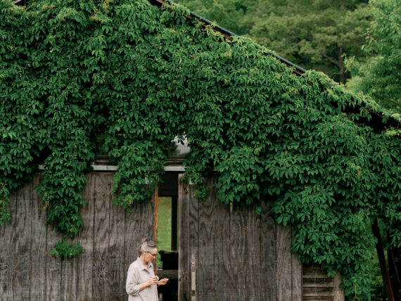 A woman stands in front of a vine-covered barn with two small dogs