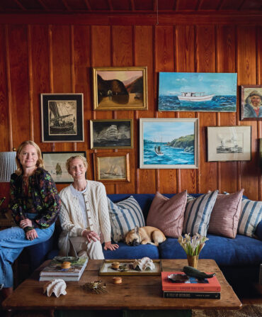 Two women sit on a couch in a wood-paneled living room.