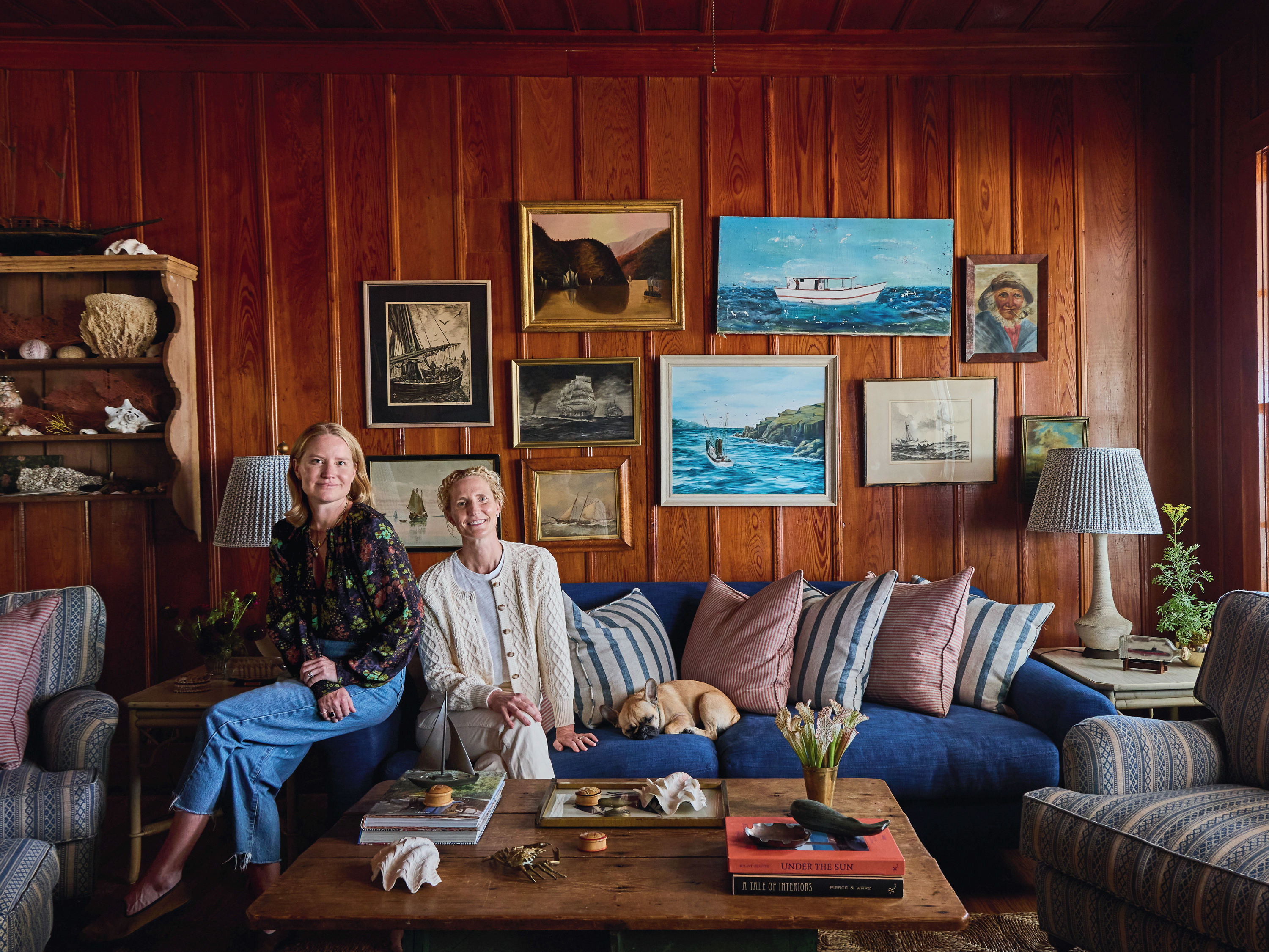 Two women sit on a couch in a wood-paneled living room.