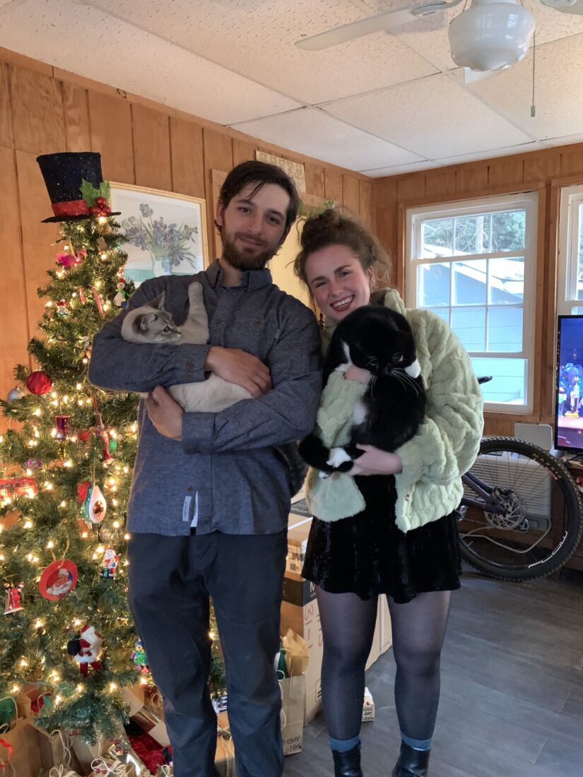 A couple smiles in front of a Christmas tree. They hold two cats