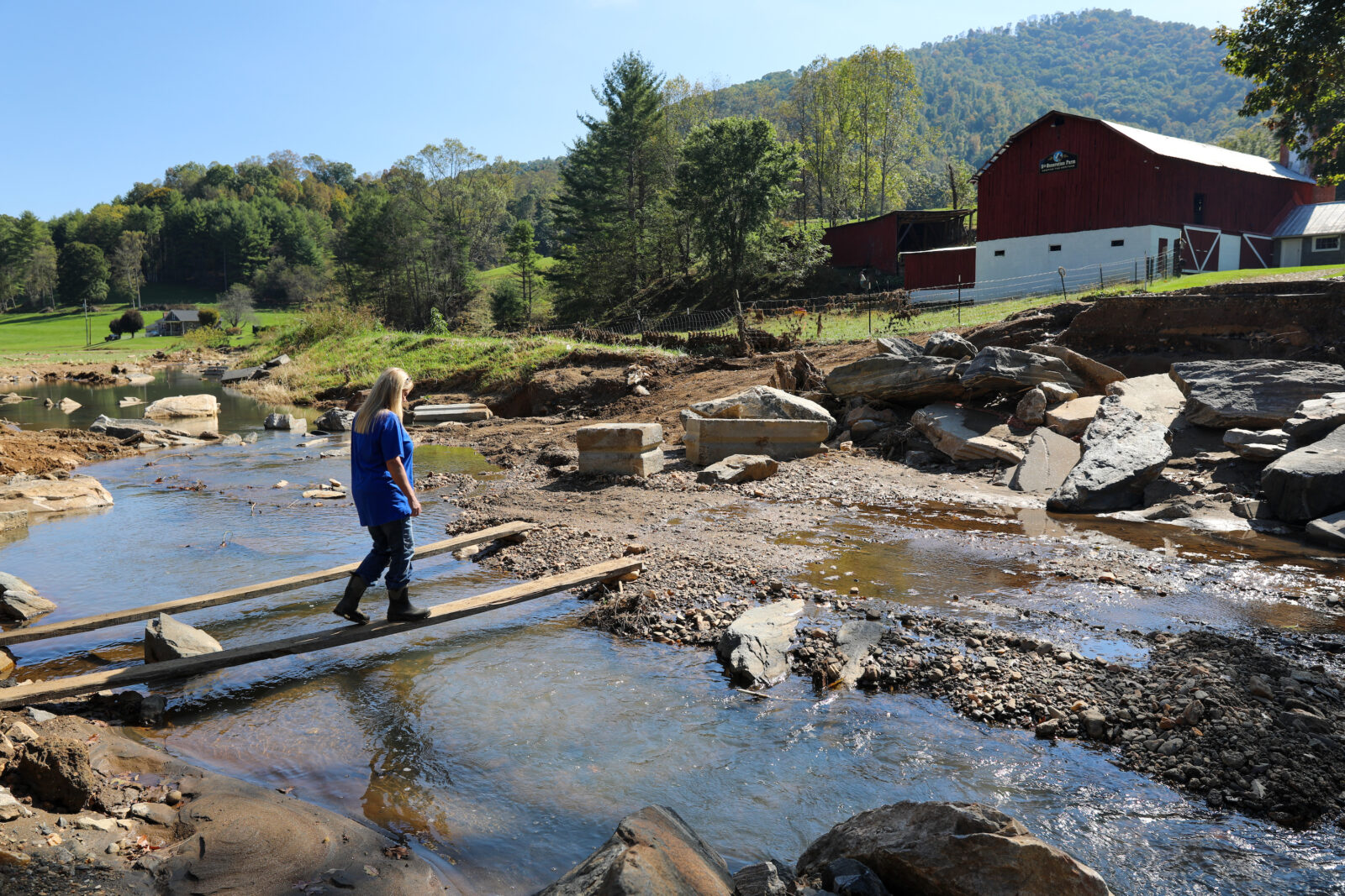 A woman walks across a plank over water toward her farm
