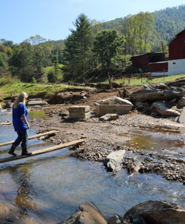 A woman walks across a plank over water toward her farm