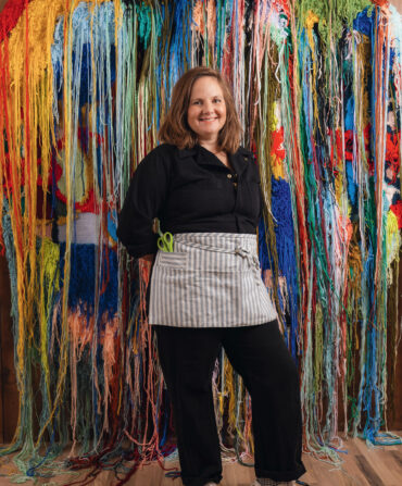 A woman stands in front of a colorful tapestry