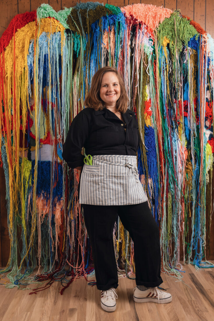 A woman stands in front of a colorful tapestry