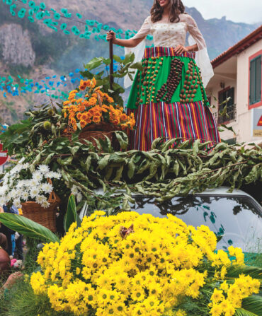 A woman in vibrant clothing stands atop a mound of flowers against a mountain background