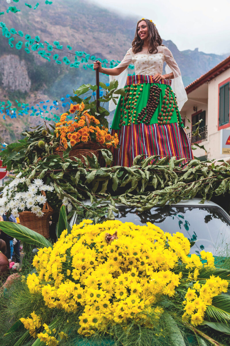 A woman in vibrant clothing stands atop a mound of flowers against a mountain background