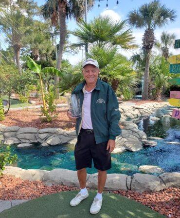 A man holds a glass trophy on a mini golf course