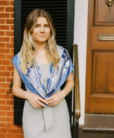 A girl stands against a brick background wearing a blue silk scarf around her neck