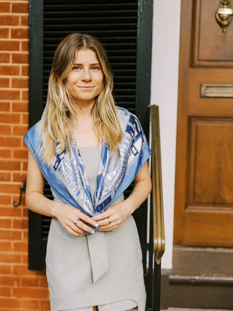 A girl stands against a brick background wearing a blue silk scarf around her neck