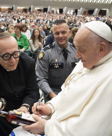 A priest and Pope Francis. Pope Francis holds bottles of bourbon and signs them