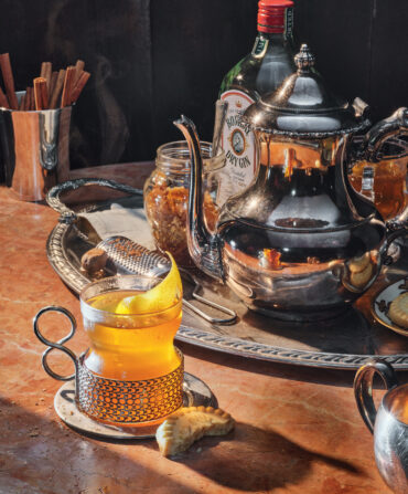 A hot cocktail in a glass sits by a half-eaten cookie and a silver platter with a silver tea pot and a bottle of gin