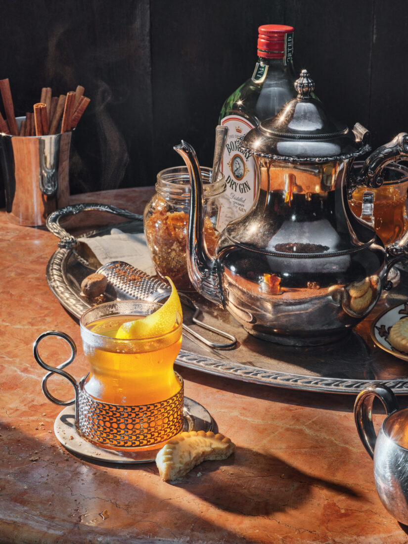 A hot cocktail in a glass sits by a half-eaten cookie and a silver platter with a silver tea pot and a bottle of gin