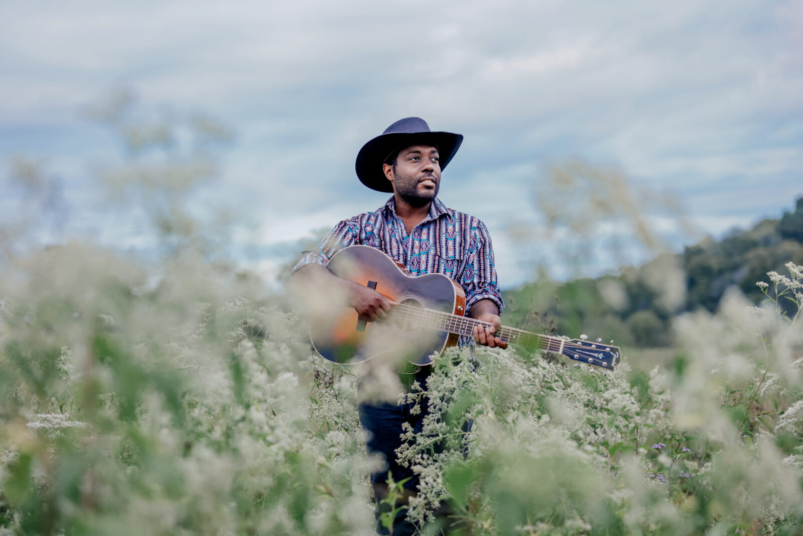 A woman with a cowboy hat and guitar stands in a field
