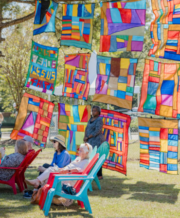 Rows of pieced together colorful quilts hang above seated people