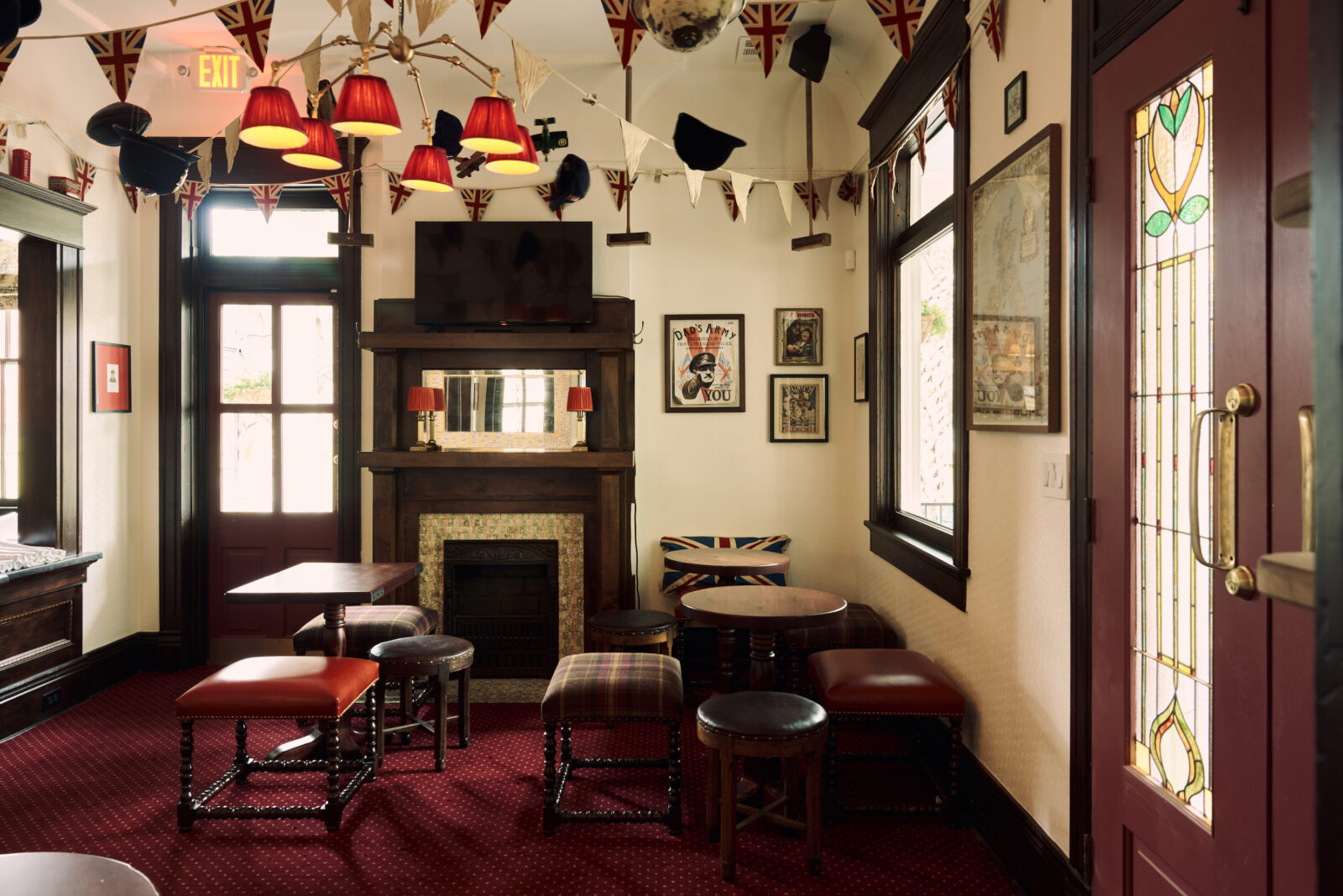 A pub room with spindle-legged furniture, flags hanging from the ceiling, and a red carpet