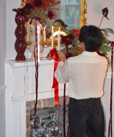 A person lights taper candles on a fireplace mantel with flowers and ribbon